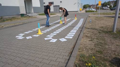 Uczestniczka gry wraz z nauczycielem na planszy do gry terenowej. Kartki z numerami ułożone na kostce brukowej.