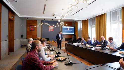 Uczestnicy spotkania siedzący po obu stronach stołu konferencyjnego, osoba prowadząca spotkanie stoi na środku sali obrad przed ekranem z prezentacją.