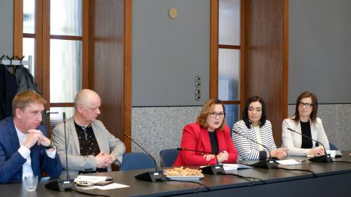 Fragment spotkania z udziałem uczestników siedzących w rzędzie przy stole konferencyjnym, jedna z osób omawia temat, pozostali notują lub słuchają.