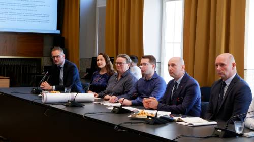 Uczestnicy spotkania siedzący w rzędzie przy stole konferencyjnym, skupieni na prezentowanych danych wyświetlanych na ekranie. Na stole widoczne dokumenty, mikrofony i materiały robocze.
