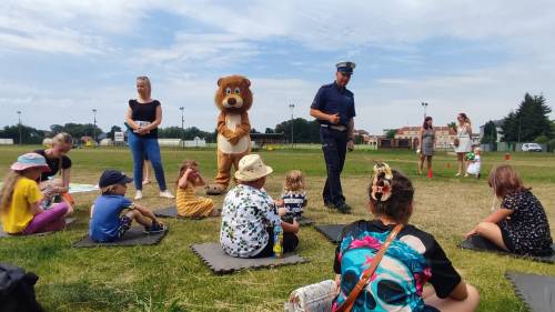 Dzieci siedzą na trawie i słuchają policjanta, na zdjęciu też Pruszek