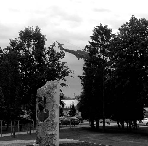 Śmigłowiec między drzewami. Pomnik ofiar Zbrodni katyńskiej na pierwszym planie - zdjęcie czarni-białe.